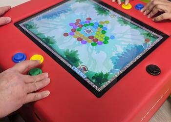 Auf dem Bild ist der Spieletisch Kuti zu sehen, an dem zwei Personen gerade etwas spielen
